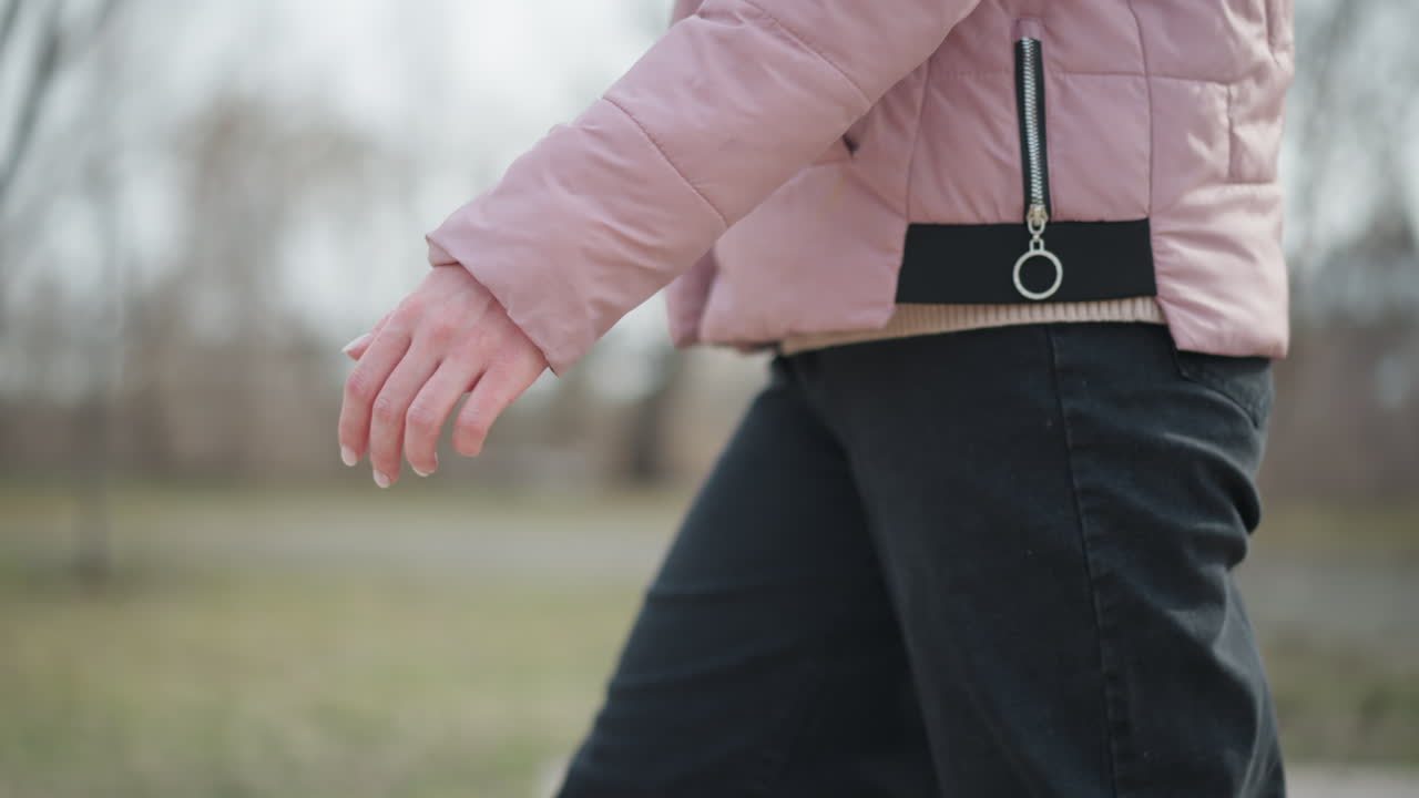 Close-up of female hand relaxed by side while walking in cold weather, wearing pink winter jacket and black pants, soft natural lighting, blurred outdoor background, focus on casual motion