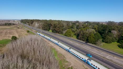 Blue-passenger-train-moving-along-tracks-beside-a-highway,-rural-landscape-on-the-outskirts-of-Buenos-Aires