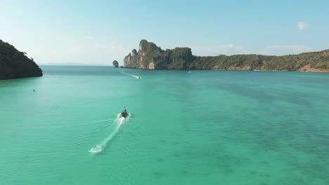 long-tail boat sailing through idyllic emerald green sea in loh dalum bay in ko phi phi don island, thailand - aerial fly-over panorama shot