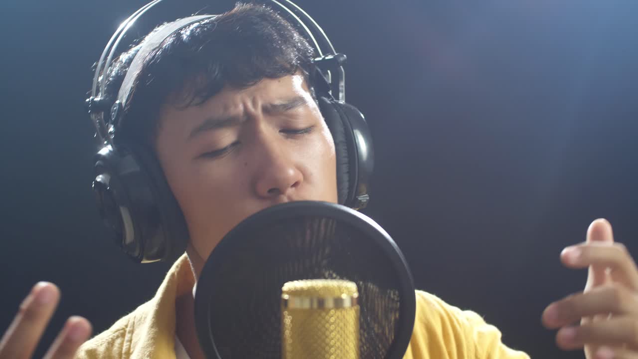 젊은 아시아 소년의 가까운 사진. 헤드폰을 들고 검은 바탕에 있는 응축기 마이크를 하고 있습니다.