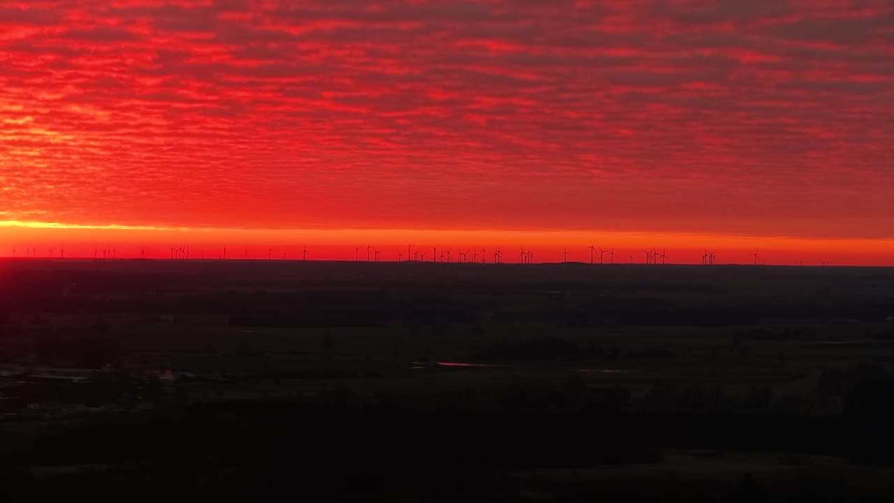 Ein atemberaubendes Drohnenvideo, das eine Luftaufnahme eines Windparks unter dramatisch rotem Himmel bei Sonnenuntergang zeigt. Die weitläufige Landschaft bietet eine ruhige, stimmungsvolle Landschaft mit einem lebendigen, feurigen Himmel.