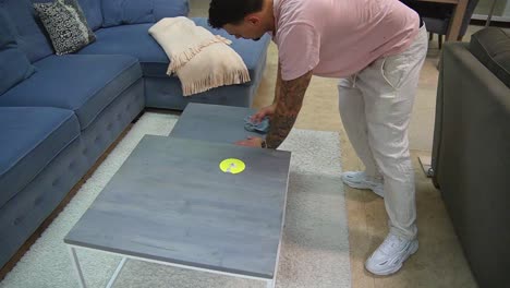 young male worker cleaning small side tables with wipe inside furniture store.