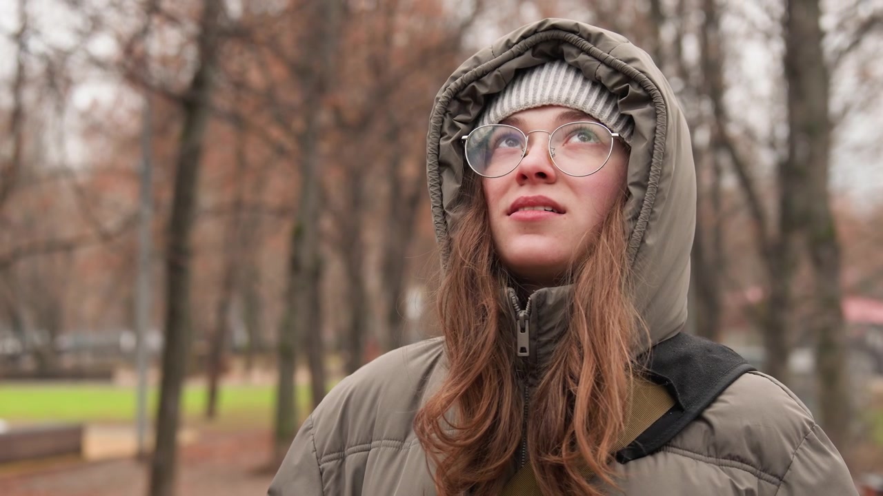 Donna bianca in un parco che indossa un cappotto con cappuccio, occhiali e uno zaino, con un sorriso gentile. Alberi autunnali spogli e cielo coperto. Parka e berretto ben avvolti. Ritratto spontaneo che cattura un'atmosfera serena, contemplativa e delicata.