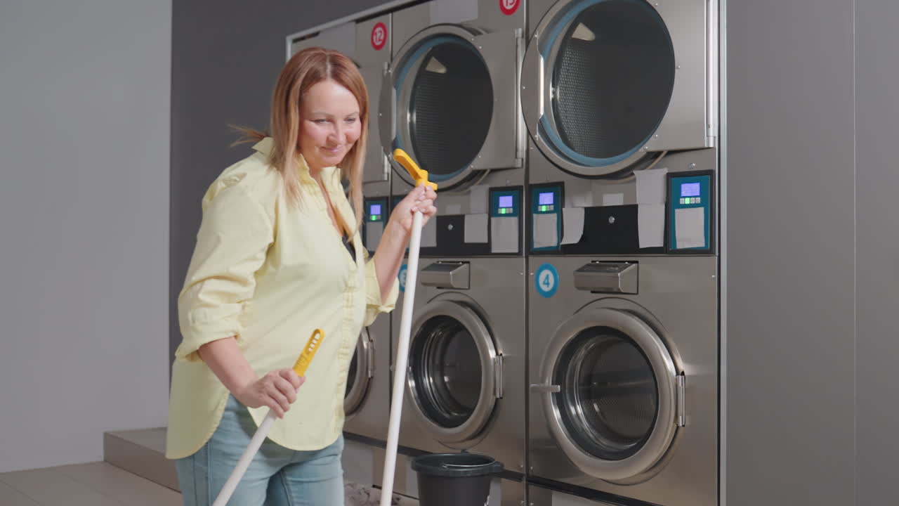 Jovial homemaker dances while sweeping near industrial washer in laundromat, holding broom stick and dust pan, cleaning lint spilled from filter drawer on floor during maintenance