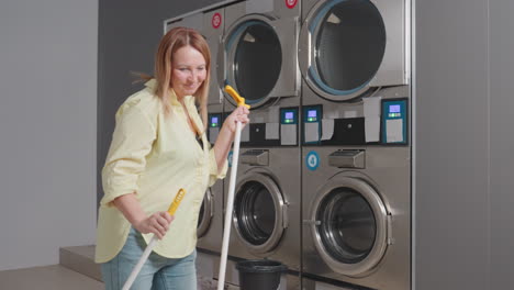 jovial homemaker dances while sweeping near industrial washer in laundromat, holding broom stick and dust pan, cleaning lint spilled from filter drawer on floor during maintenance