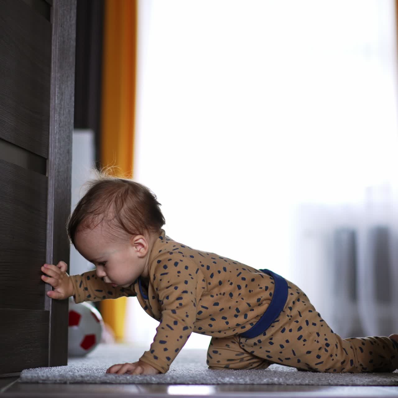 Little baby in brown romper crawls up to the doorway. Beautiful boy pushes the doors and falls on the floor