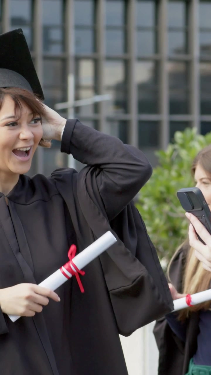 Vertical video: Panning cam grads in gowns raising diploma fixing caps by quad using phone for pics
