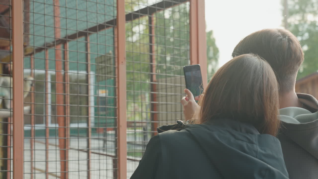 two white teens filming monkey enclosure, capturing closeup shots through metal mesh, handheld phone recording candid curiosity, educational signage, daylight outing and playful interaction