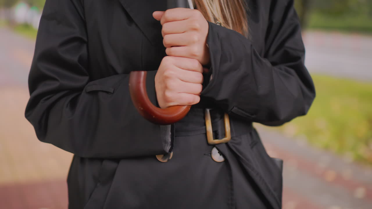 Close-up of woman in black trench coat with hood holding umbrella tightly on rainy autumn day, capturing detail of hands, clothing, wood handle, and moody seasonal atmosphere with blurred background