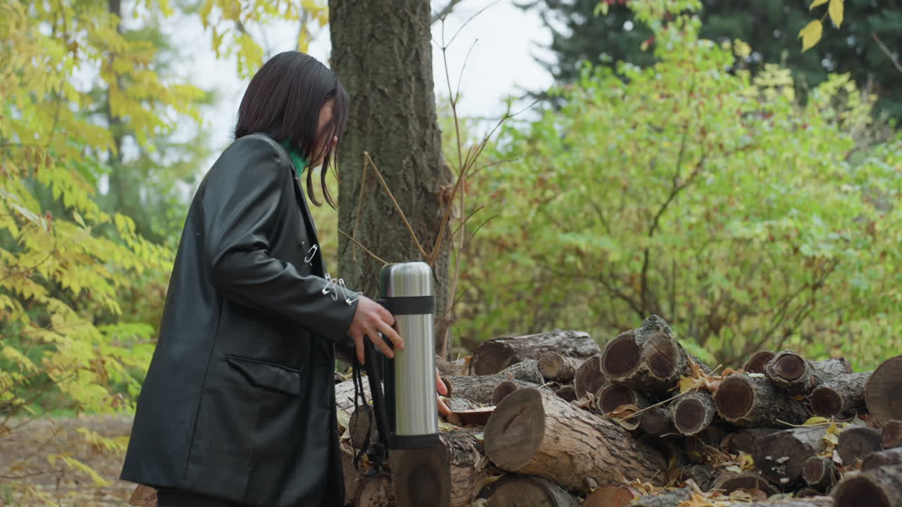 Black woman in leather jacket pauses to pour coffee from metal thermos onto stacked logs in autumn forest, braided hair with green scarf, candid side profile, quiet contemplative mood, soft natural
