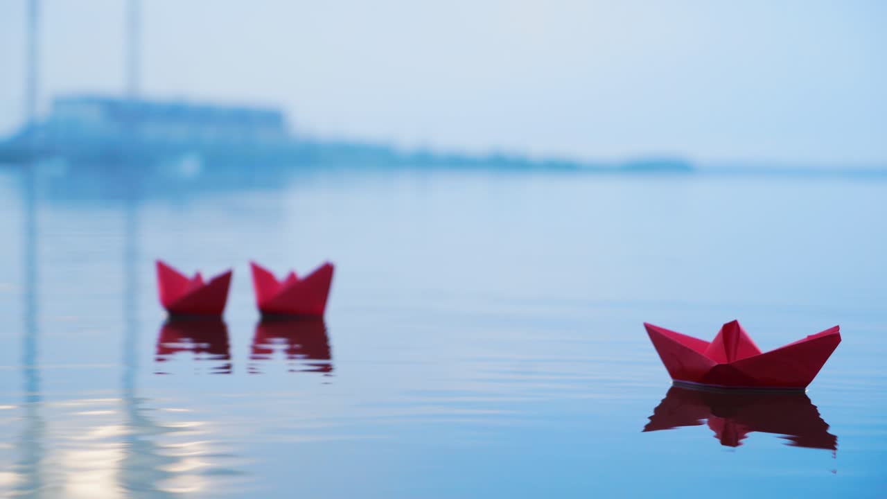 Red paper origami boats floating on the clear water with their reflection on blurred background. Three origami boats swimming on the blue lake. Close-up