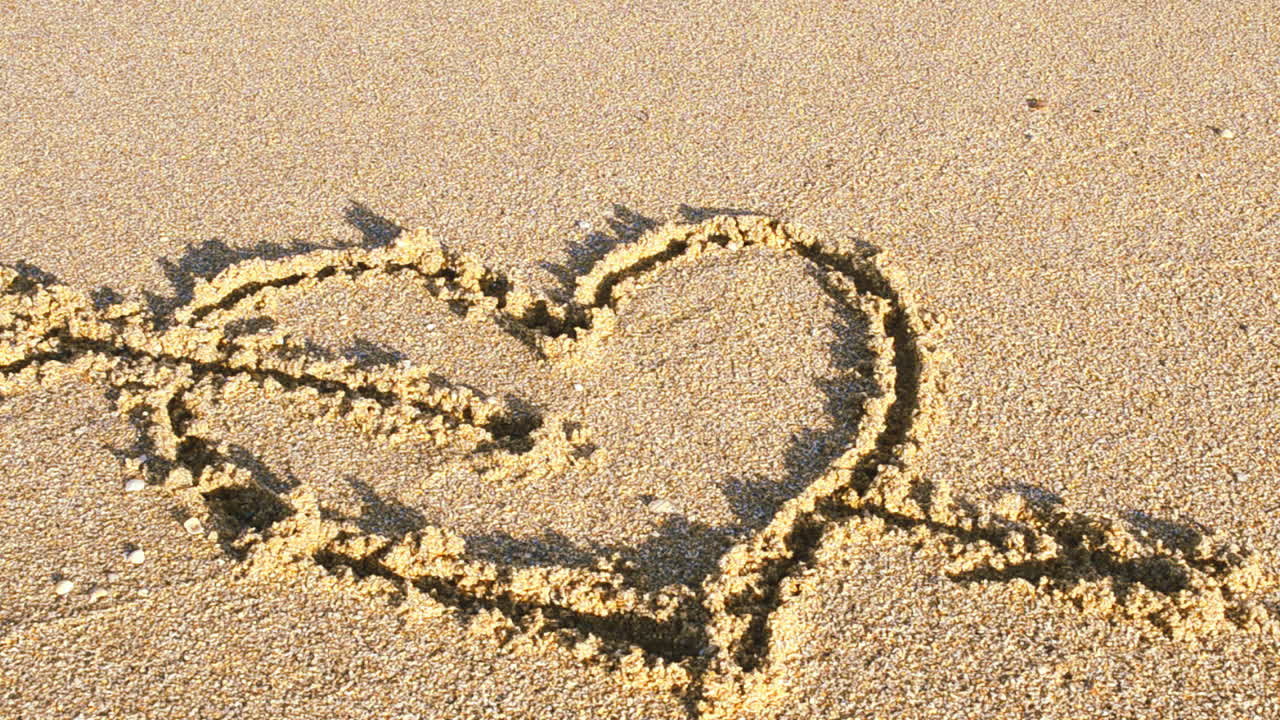 Un cuore disegnato sulla sabbia. Il cuore viene cancellato da un'onda marina. Riprese sulla spiaggia.