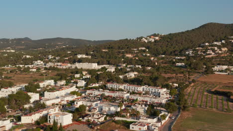 Aerial-slow-motion-view-of-beautiful-small-white-houses-in-a-colony-with-parked-cars-and-on-street-surrounded-by-greenery-and-mountains-during-evening-in-the-beautiful-city-Ibiza-in-Spain