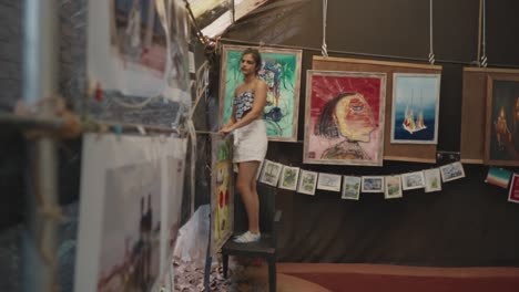 A-full-body-shot-captures-an-Indian-woman-creatively-using-a-chair-to-reach-greater-heights-while-hanging-artwork-in-an-art-gallery,-displaying-her-resourcefulness-and-determination