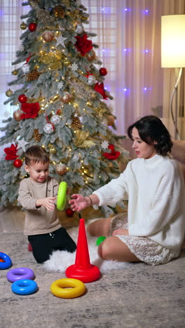 happy family of mother and son play near the christmas tree. mom and baby play with a toy pyramid. vertical video.