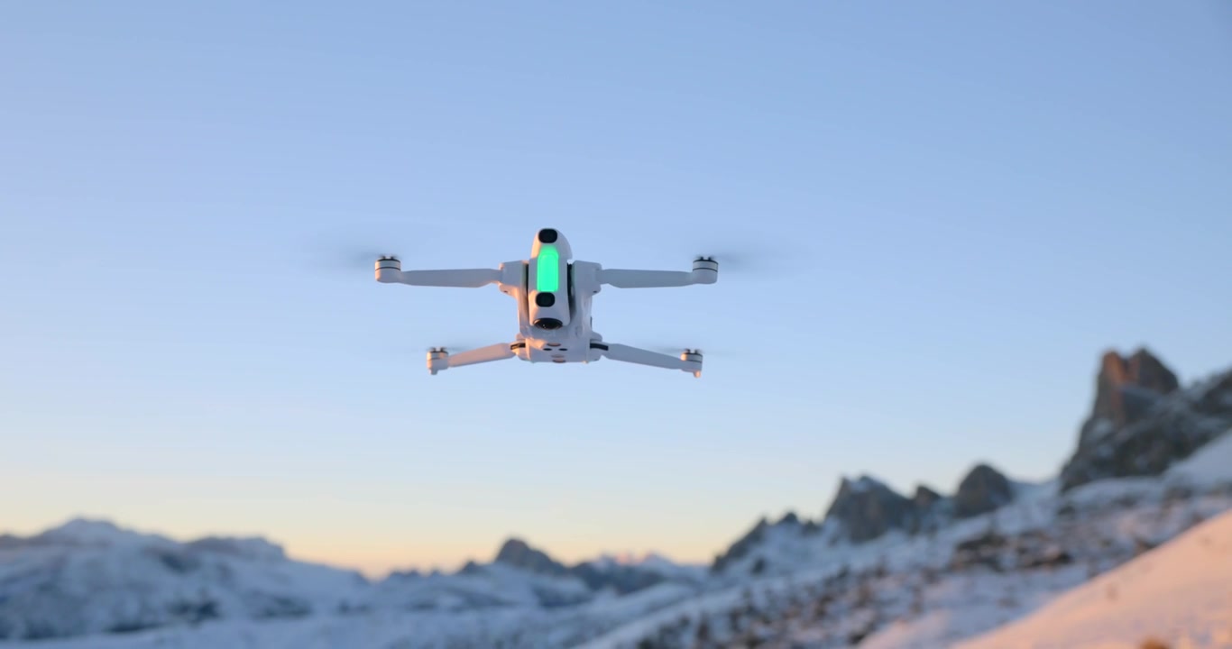 Vista dal basso di un drone quadricottero bianco che si libra in cielo sereno sopra cime montuose innevate al tramonto.