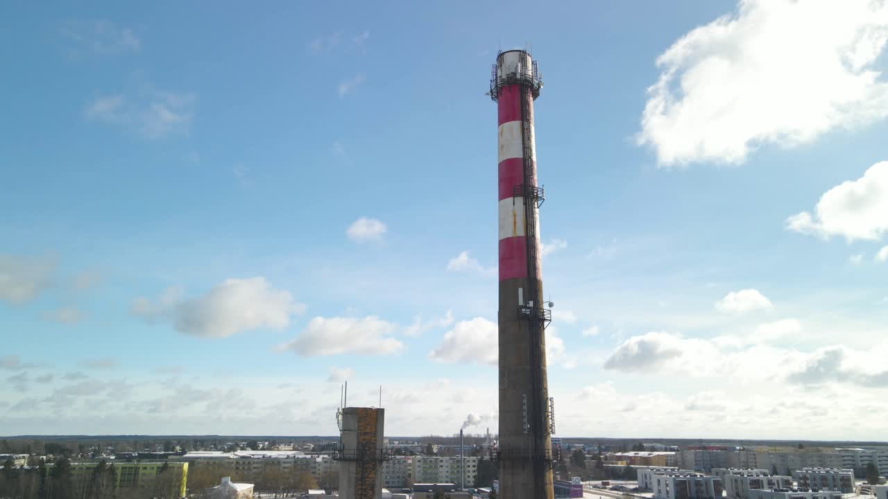 Aerial drone view footage that is circling and spinning around a large smoking CHP plant tower that produces energy and thermal heat and warmth. The concrete tower has white and red stripes, sunny day