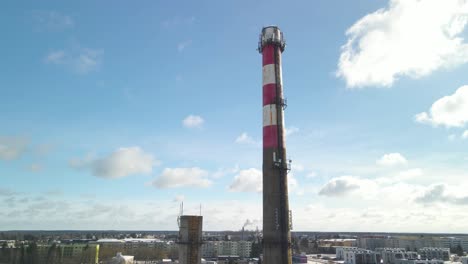 aerial drone view footage that is circling and spinning around a large smoking chp plant tower that produces energy and thermal heat and warmth. the concrete tower has white and red stripes, sunny day