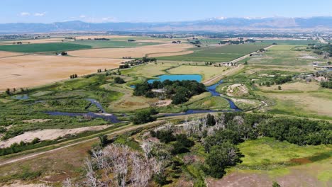 Panoramic-video-of-picturesque-pastures,-river-canals,-and-a-tranquil-blue-lake-nestled-in-a-valley-with-majestic-mountains-on-the-distant-horizon,-under-a-vast-blue-sky,-in-Colorado