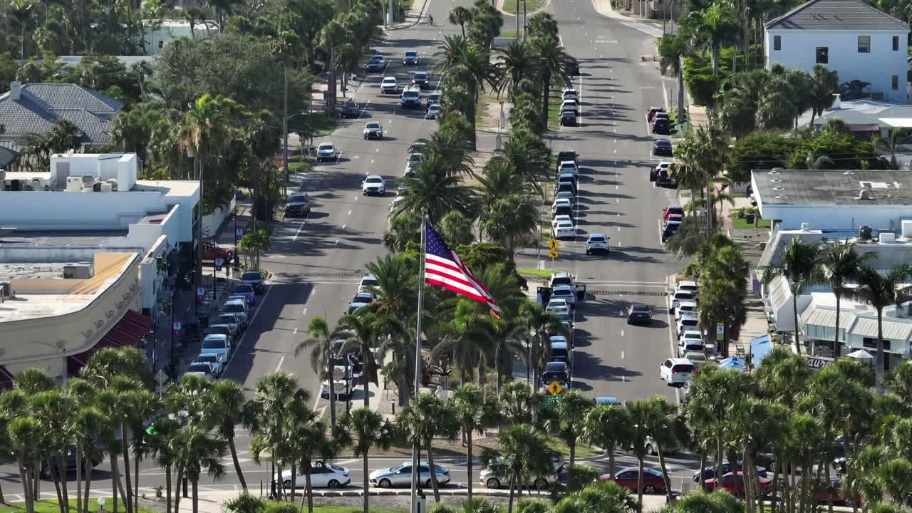 American flag waving on busy street with cars of Sarasota, Florida. Aerial tilt up wide shot. Palm tree avenue with river and luxury houses in background. Sunny summer day in United States.