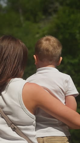 caring mother holds little son showing picturesque waterfall in wild nature park. woman with toddler boy near high water cascade on weekend vacation