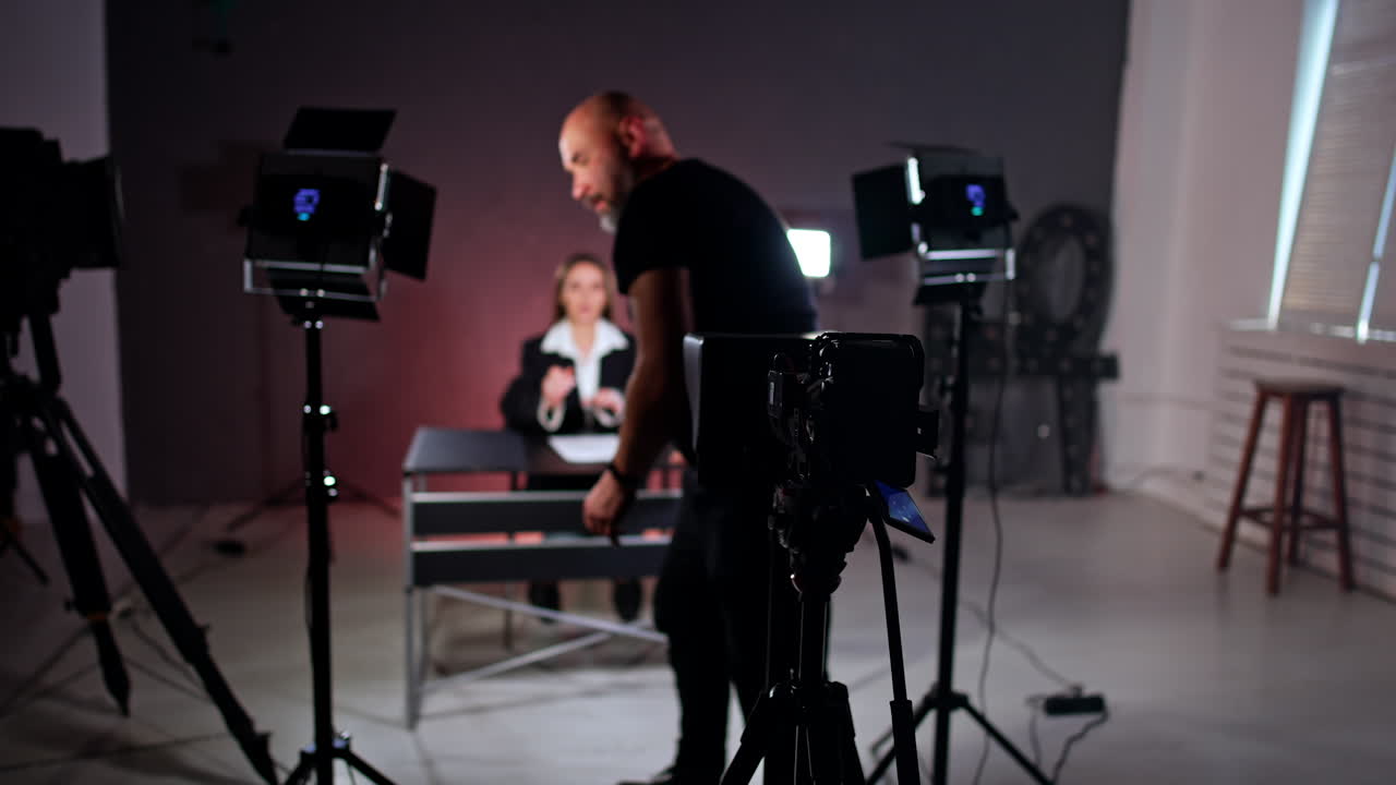 Cameraman leaves his place behind the camera and approaches a lady sitting at desk. Photographer fixes the objects at the desk for better look. Blogging concept.