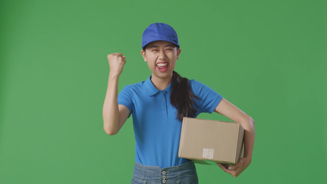 Asian Female Courier In Blue Uniform Celebrating Succeed Delivering A Carton On Green Screen Background In The Studio