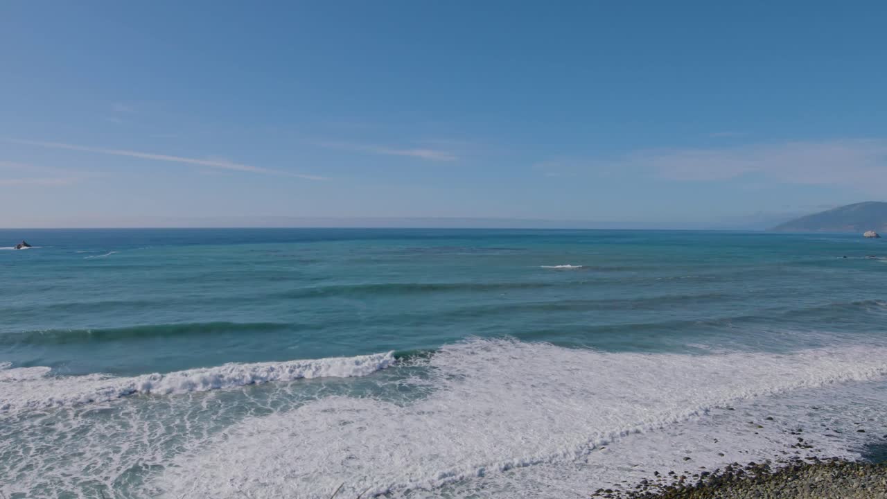 Wide aerial shot of rolling waves crashing gently onto a rocky shoreline beneath clear skies on California’s Big Sur coast.