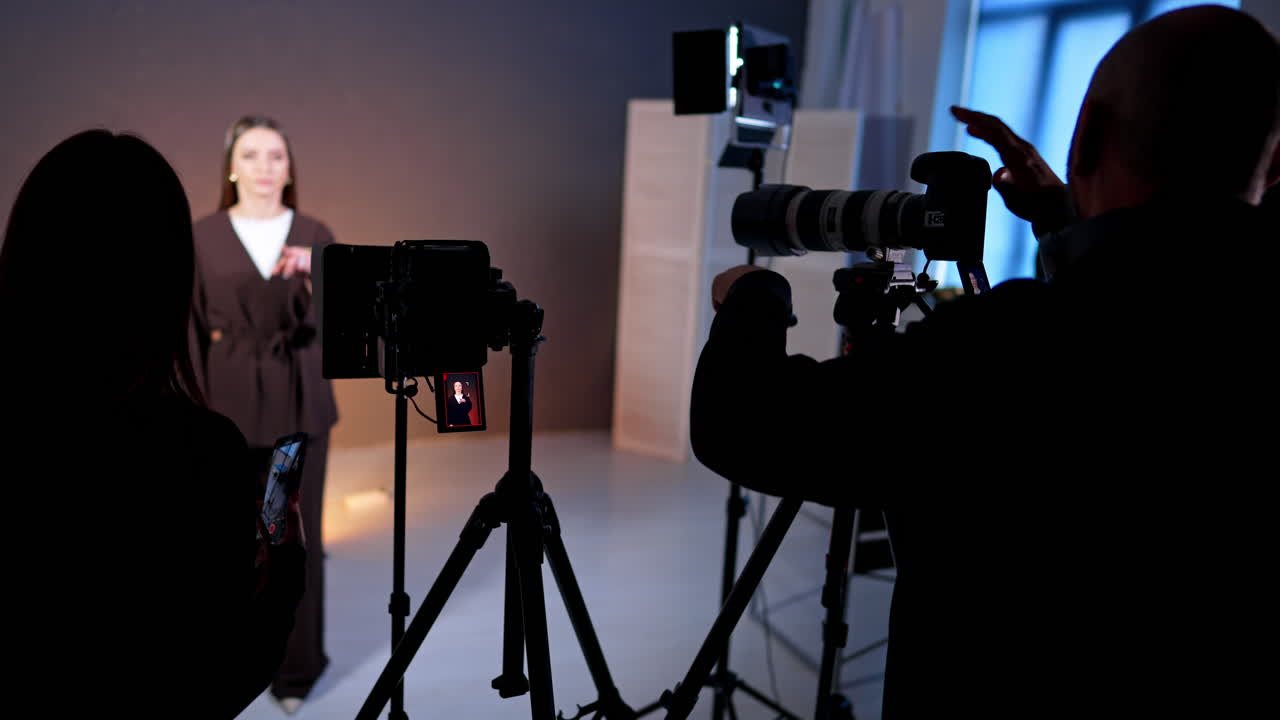 Silhouette of video crew filming female presenter in dark outfit. Crew members operate cameras in dark studio while woman in brown suit speaks in focused spotlight