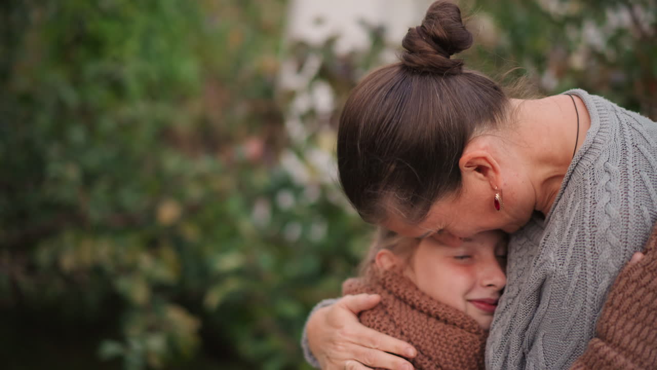 Madre che abbraccia la figlia all'aperto in giardino, con un caldo maglione lavorato a maglia e una sciarpa, un tenero bacio sulla fronte, primo piano con bokeh cinematografico, toni morbidi e caldi, un abbraccio emotivo e protettivo e un sorriso gentile che trasmettono l'idea di famiglia.