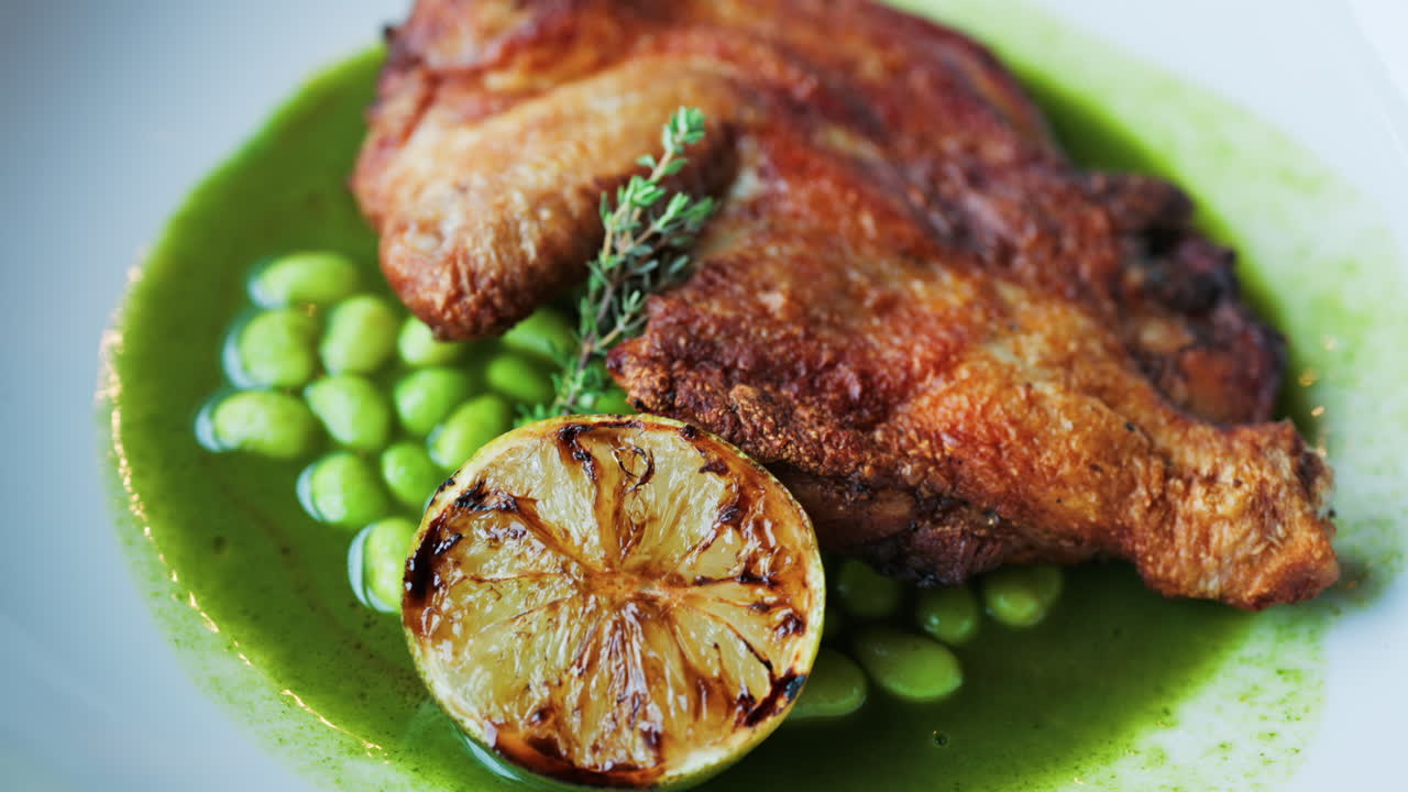 Close-up of a gourmet roasted chicken thigh served with edamame beans, a drizzle of herb-infused green sauce, and a charred lime half