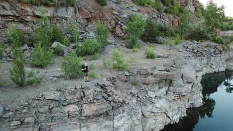young tourist man standing on steep slope with backpack over the water. camper observing beautiful view of canyon with green trees and bushes and their reflection in the water. aerial view