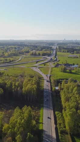 Vertical drone panning view captures Katlakalns highway merging into multiple lanes through green countryside with forest belts, farmhouses, and Riga skyline far in the background