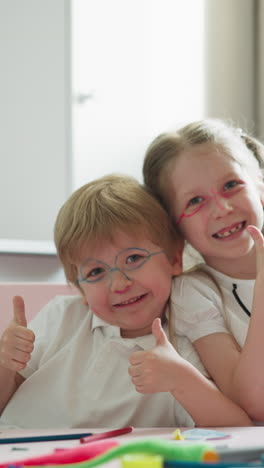 brother and sister with painted glasses bump fists friendly sitting at desk with books and felt-tip pens. little siblings show thumbs-up smiling joyfully