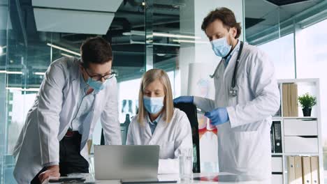 caucasian senior female doctor in medical mask sitting in hospital, typing on laptop and speaking with male colleagues at work