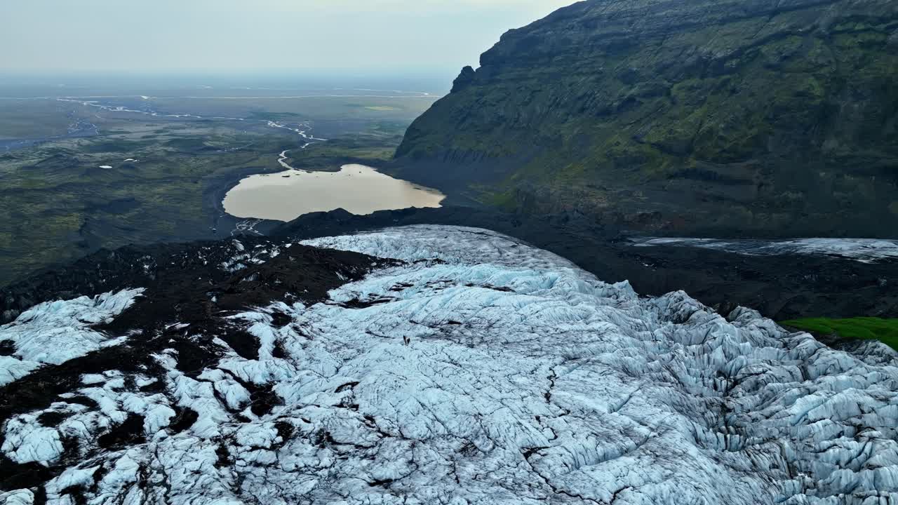 A wide establishing aerial shot revealing a glacier cascading toward a pale lake beneath steep volcanic mountains, showcasing rugged ice textures and a vast Icelandic landscape