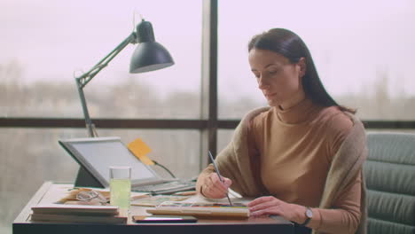 una mujer joven con un pincel y pinturas elige una sombra adecuada para el diseño del logotipo o las paredes. elección de color y sombra para el diseño futuro. una mujer colorista en una oficina con grandes ventanas dibuja con un pintal y pinta en un cuaderno.