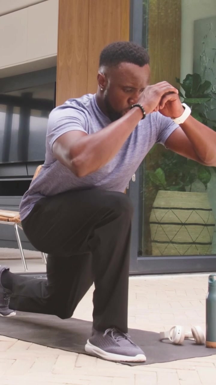 Vertical video: Glancing at watch man tracking reps doing lunges on patio in gray tee with mat