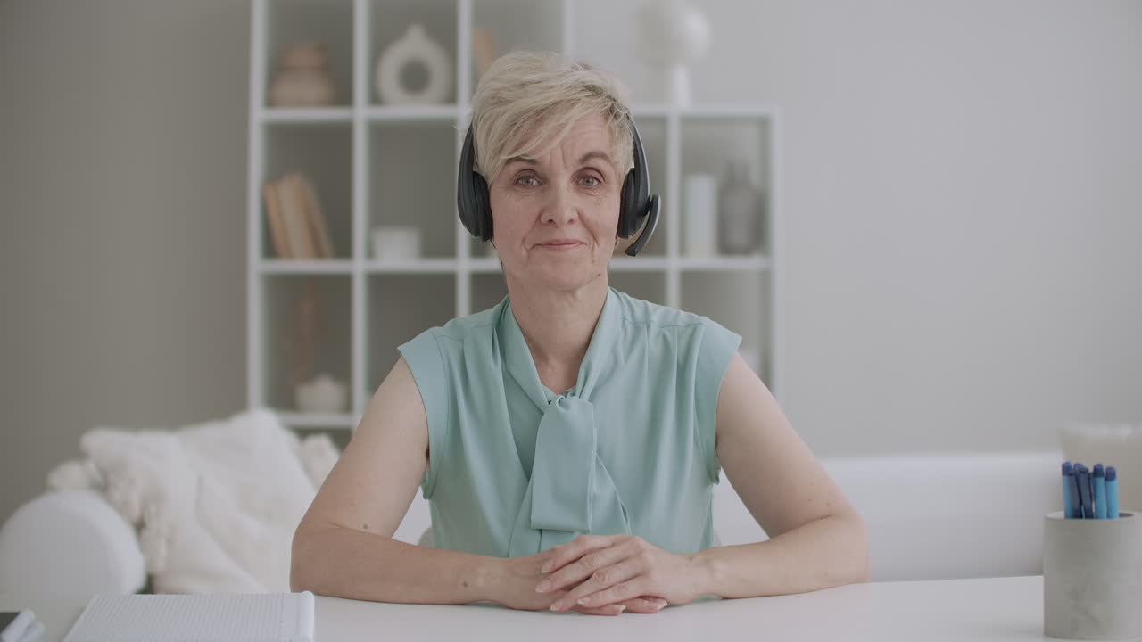 mujer de mediana edad amable y amigable está escuchando con auriculares y asintiendo sentado en la mesa y mirando a la cámara