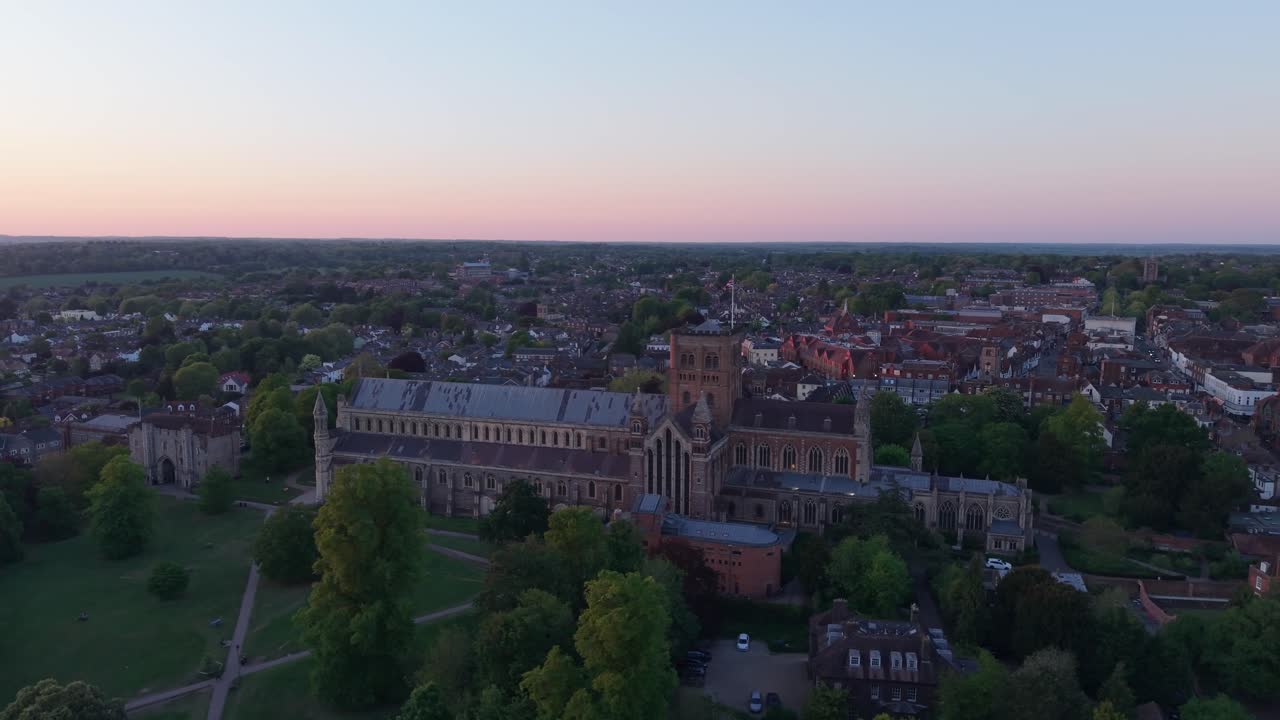 A cinematic drone orbits St Albans cityscape, highlighting the majestic cathedral against a stunning blue-orange horizon, capturing the historic urban landscape in warm light