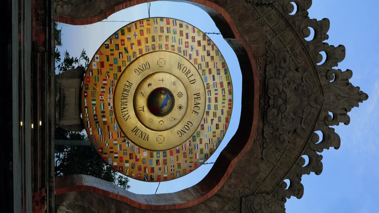 Vertcial Stunning parallax shot of the World Peace Gong in Bali adorned with golden peace signs and national flags, against a backdrop of palm trees, the moon and a vibrant blue sky