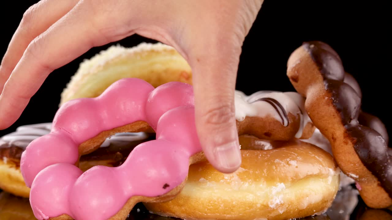 Hand picking pink glazed donut from assorted stack on reflective surface under bright studio lighting