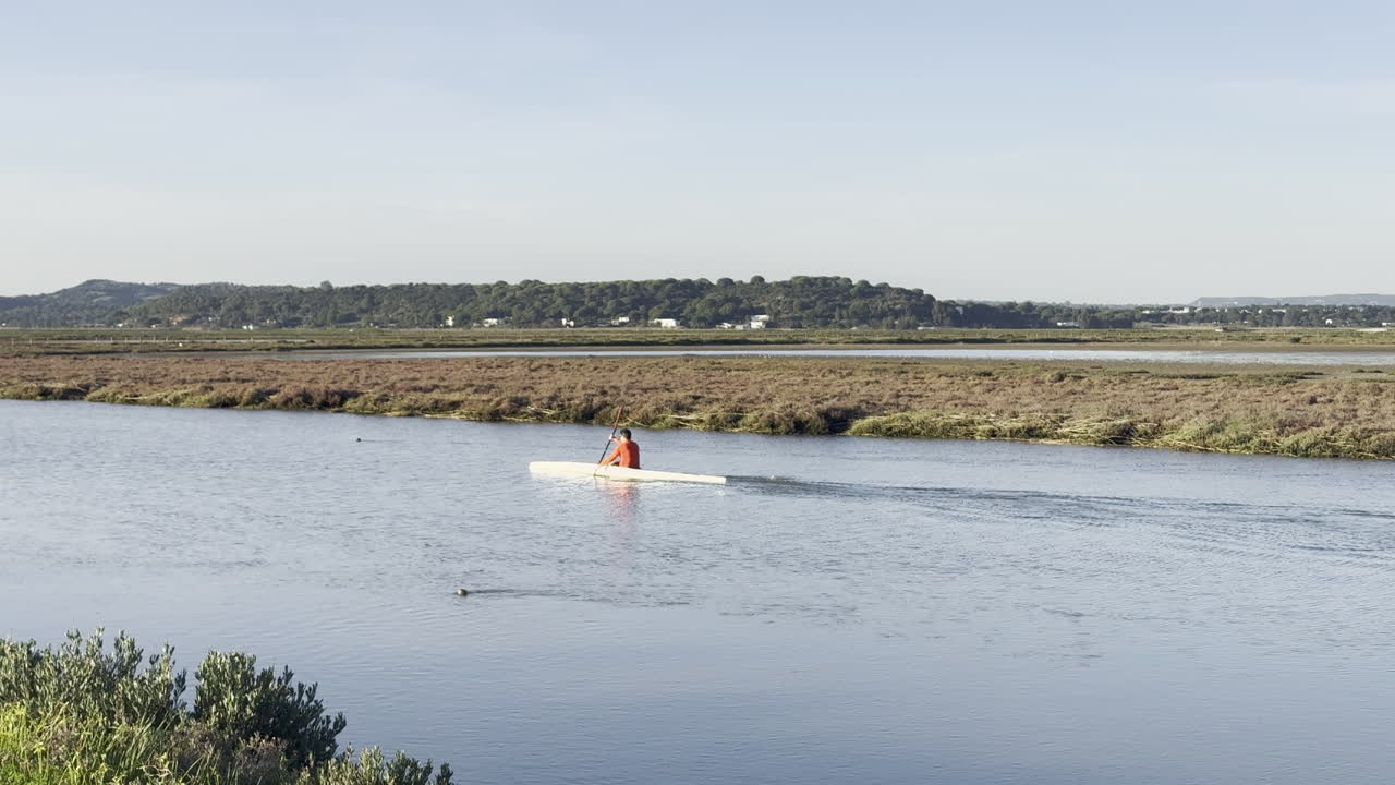 Person kayaking on calm waters with natural wetlands and distant hills under clear sky