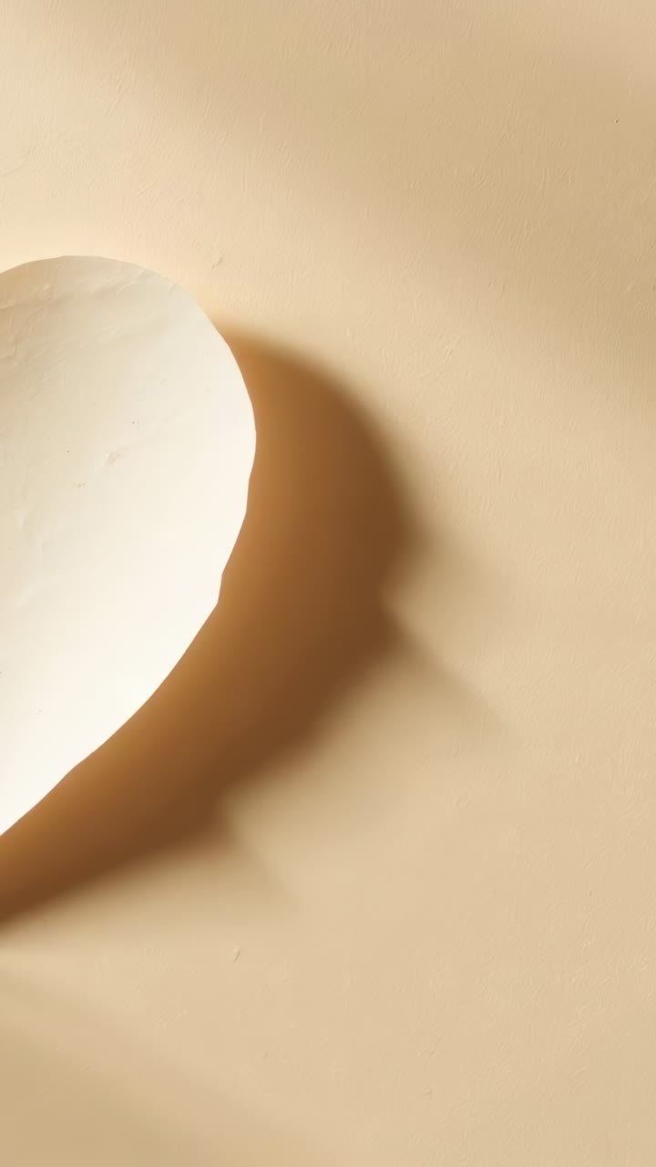 Vertical video: Sliding white heart across beige tabletop by frame shift casting shadow, copy space