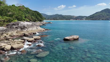 drone footage captures the serene coastline of phuket, thailand, showcasing clear waters, rocky shores, and lush greenery under bright sunlight