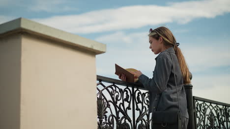 hermosa mujer con ropa gris descansa su mano en una valla de hierro mientras hojea un libro en su mano, el viento sopla suavemente su cabello, y su ropa aleta suavemente
