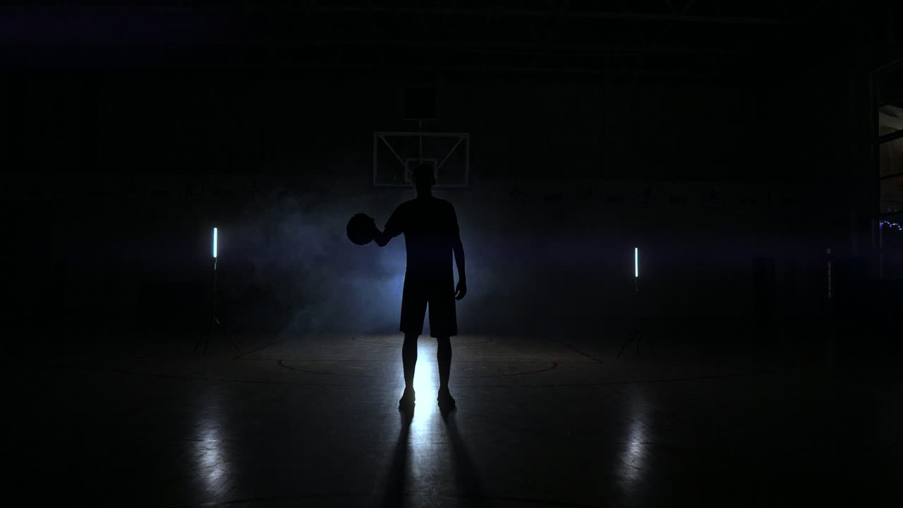 el jugador de baloncesto golpea la pelota a la luz de las lámparas que brillan detrás de las gradas en la duma y golpea la pelota en el suelo en la sala de baloncesto en cámara lenta