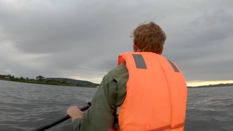 un primer plano desde atrás de un hombre con un chaleco salvavidas brillante remando en un kayak en el lago victoria en la madrugada del amanecer