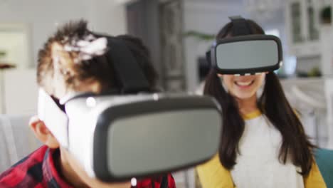 happy asian brother and sister at home, sitting on couch in living room having fun using vr headsets