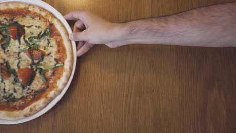 person sliding a fresh pizza across a wooden restaurant table with only a hand and arm visible